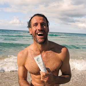 A laughing man standing near beach waves is holding a Blue Healer Daily Relief cream tube.